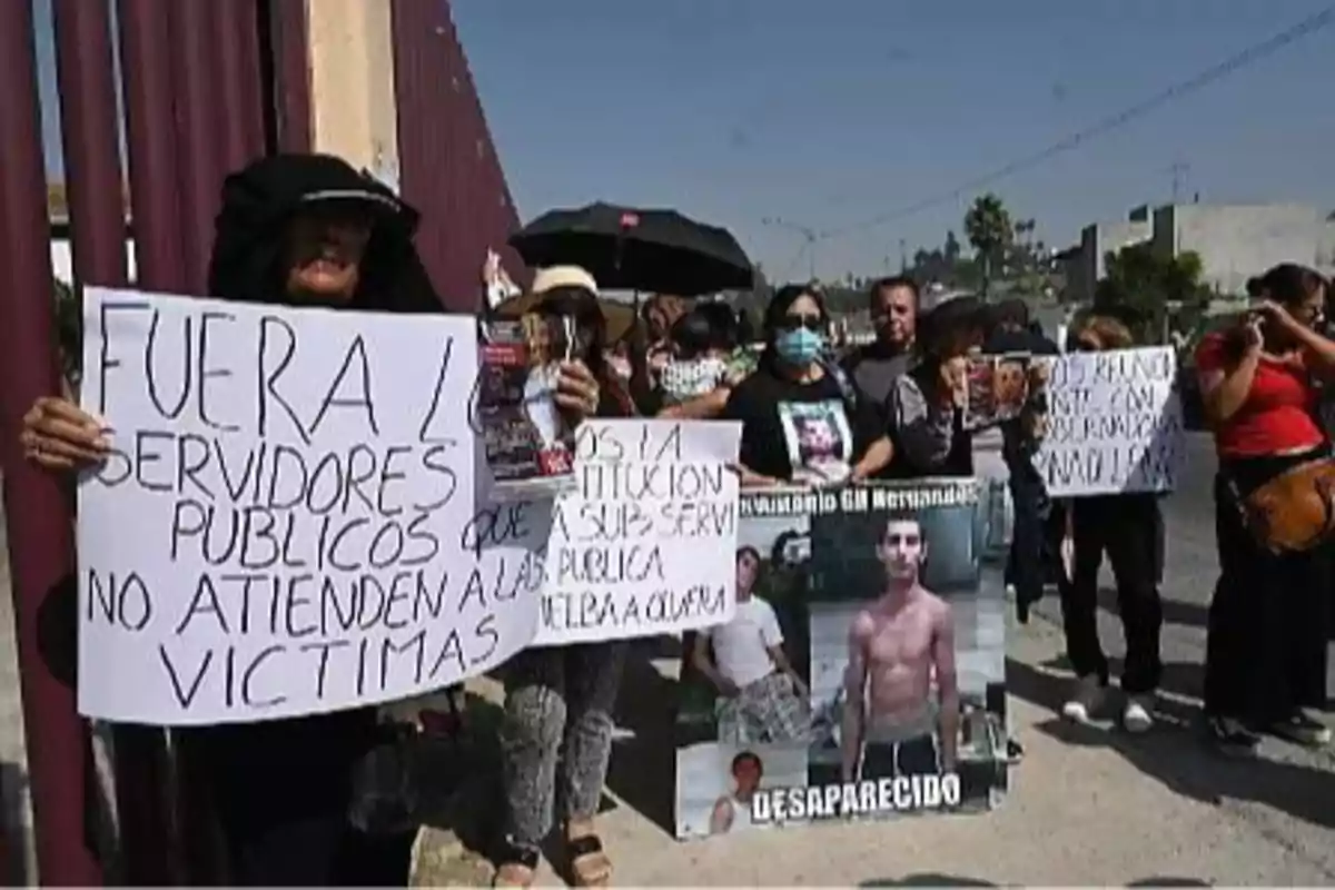 Un grupo de personas sostiene carteles de protesta en una calle, expresando descontento con los servidores públicos y pidiendo atención a las víctimas, mientras una de las pancartas muestra la foto de una persona desaparecida.