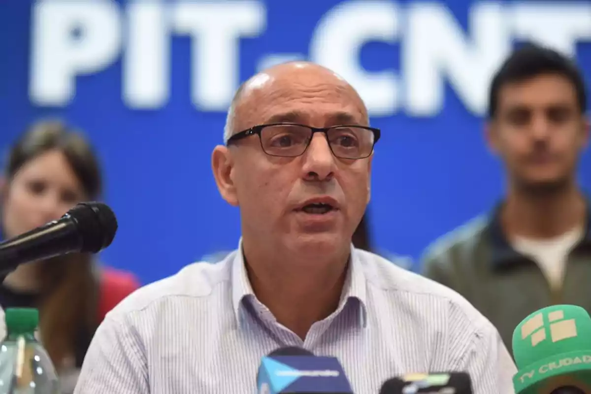 Un hombre con gafas habla frente a varios micrófonos en una conferencia de prensa con un fondo azul que tiene letras blancas.