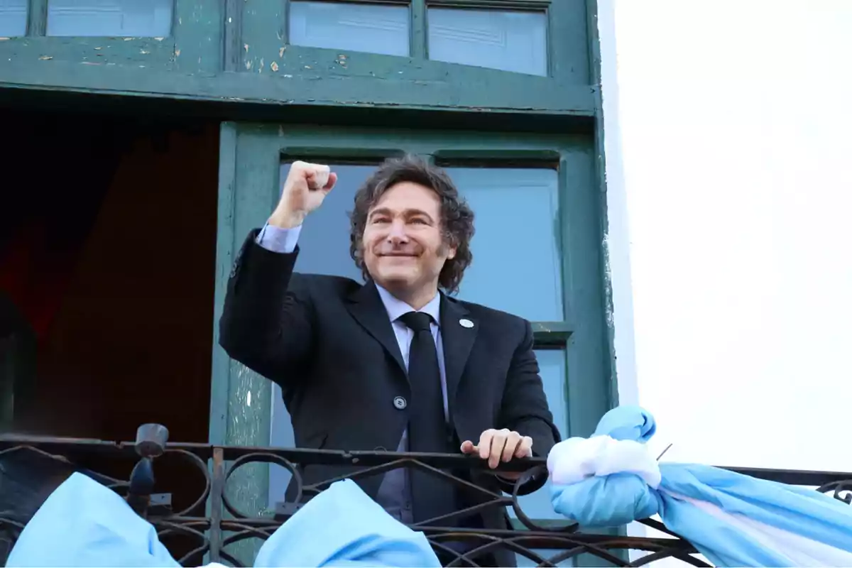 Hombre de pie en un balcón levantando el puño en señal de celebración con una bandera azul y blanca decorando la barandilla.