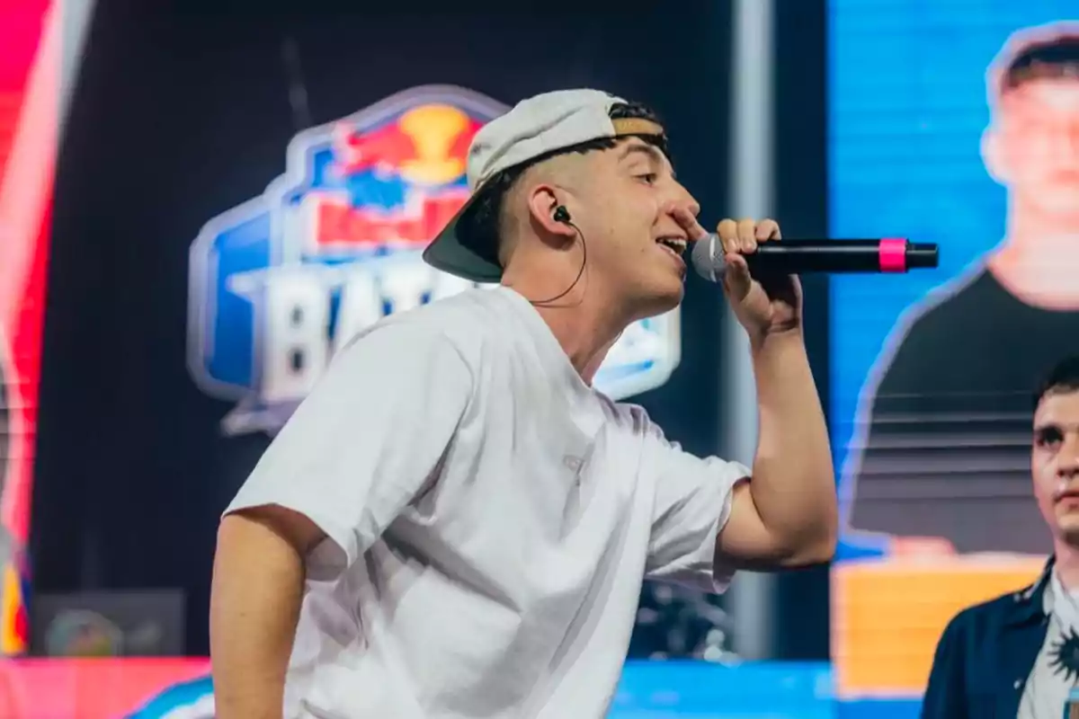 Un joven con gorra al revés y camiseta blanca sostiene un micrófono mientras canta en un escenario con un logotipo de Red Bull Batalla de fondo.