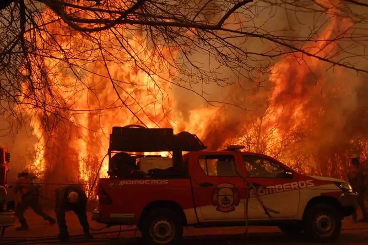 Camioneta de bomberos frente a un incendio forestal con llamas intensas y humo denso mientras los bomberos trabajan para controlar el fuego.