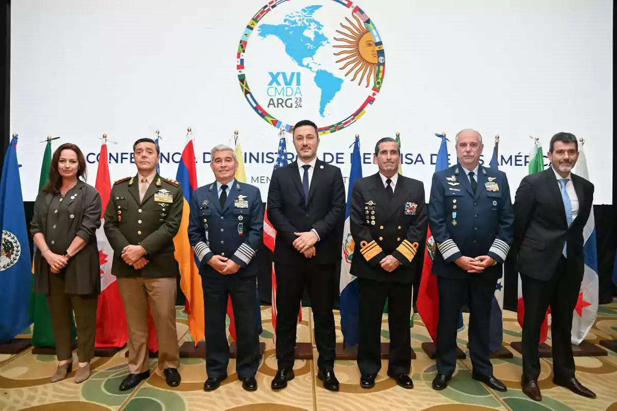 Un grupo de personas en uniforme militar y traje formal posan frente a un fondo con un logotipo de la Conferencia de Ministros de Defensa de las Américas, rodeados de banderas de varios países.