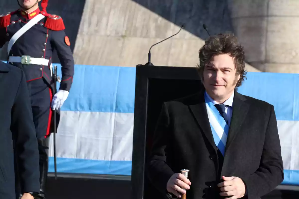 Un hombre con traje oscuro y corbata azul claro sostiene un bastón frente a una bandera argentina mientras un guardia con uniforme ceremonial está de pie al fondo.