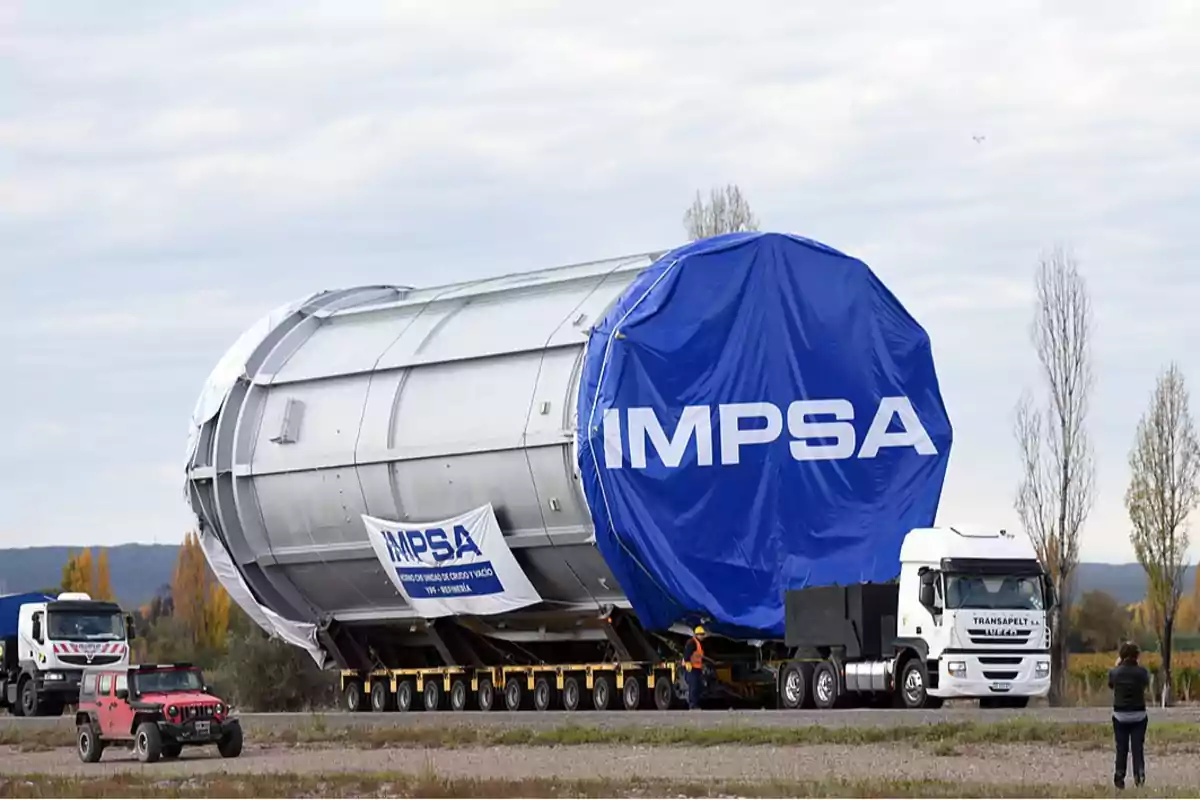 Un camión transporta un gran cilindro metálico cubierto con una lona azul que tiene el logo de IMPSA, mientras un vehículo todoterreno y otro camión lo acompañan en una carretera rodeada de árboles.