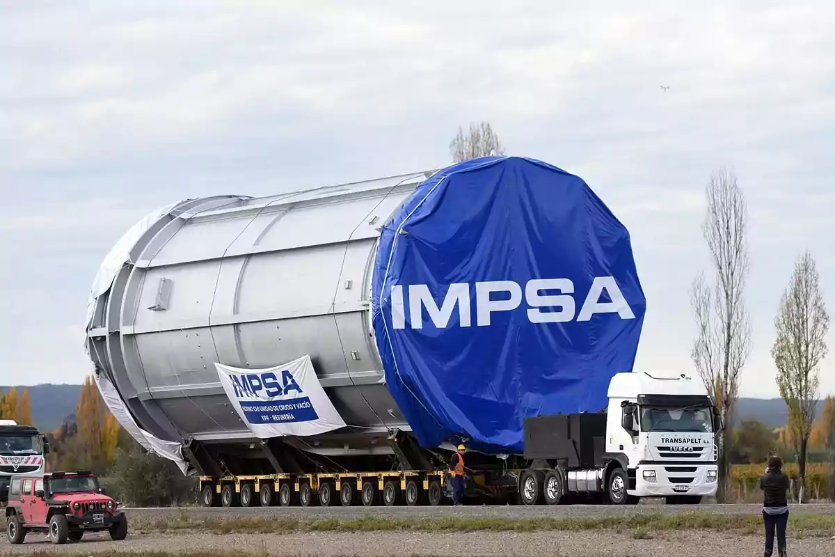 Un camión transporta un gran cilindro metálico cubierto con una lona azul que tiene el logo de IMPSA en una carretera, mientras una persona observa y toma fotos.