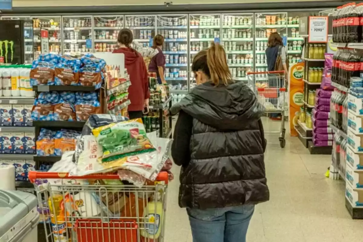 Una mujer con un carrito lleno de productos camina por el pasillo de un supermercado mientras otras personas también hacen sus compras.