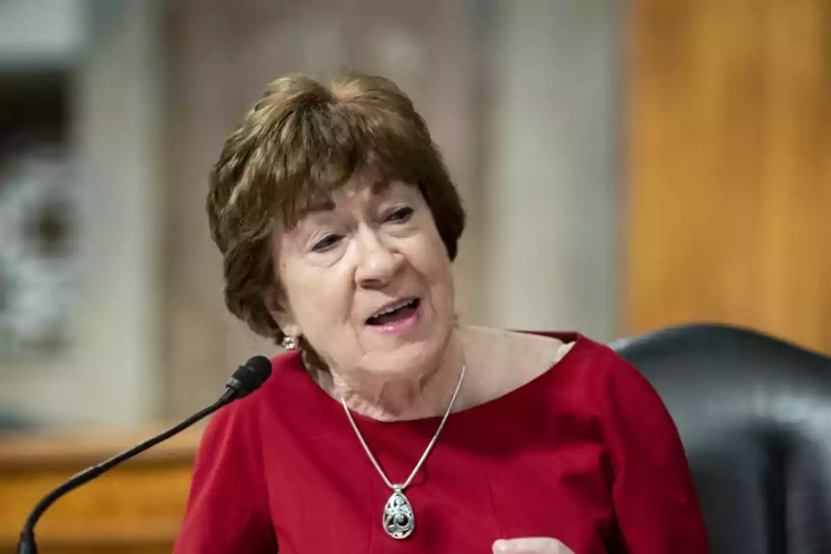 A woman with short brown hair, wearing a red sweater and a necklace, speaks in front of a microphone in a formal setting.