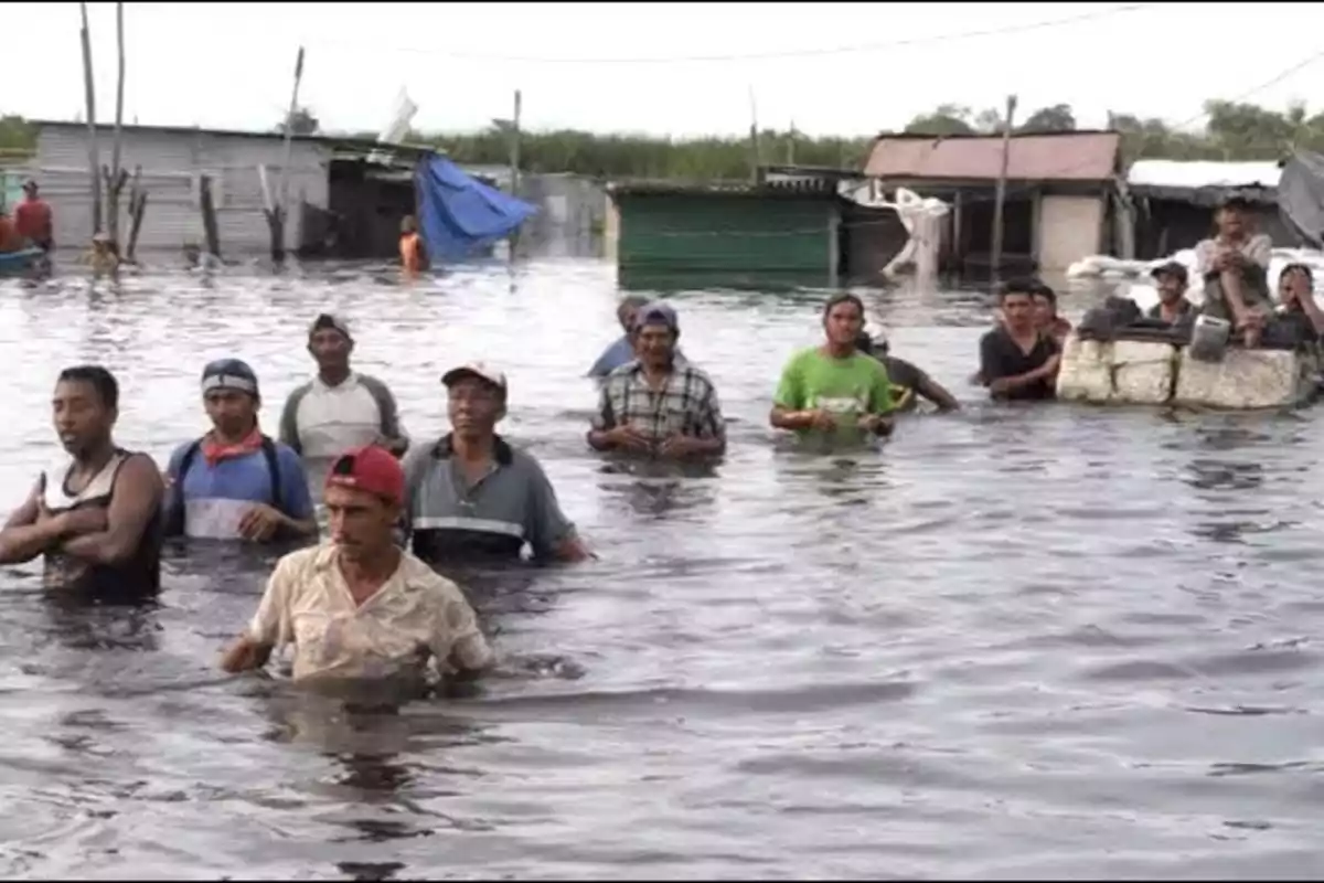 A group of people walks through a waist-deep flooded area, with houses and structures in the background partially submerged.