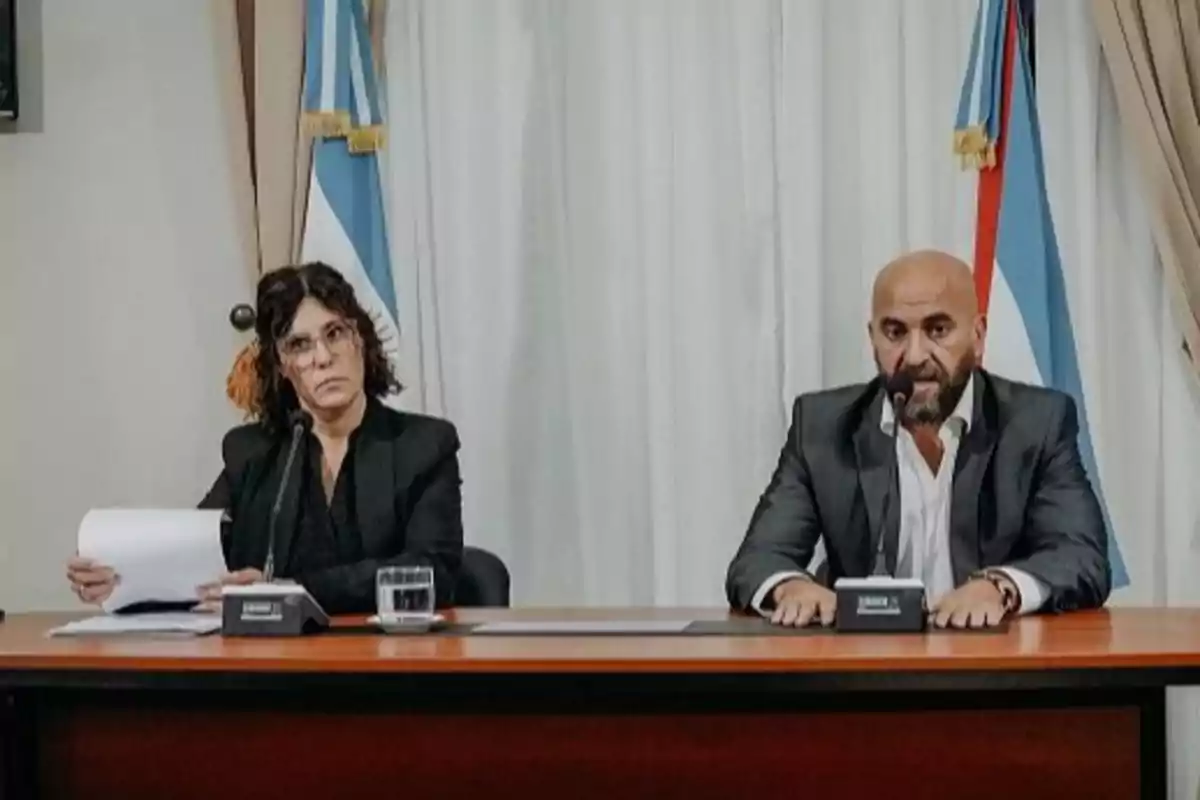 Dos personas sentadas en una mesa con micrófonos frente a banderas y cortinas.