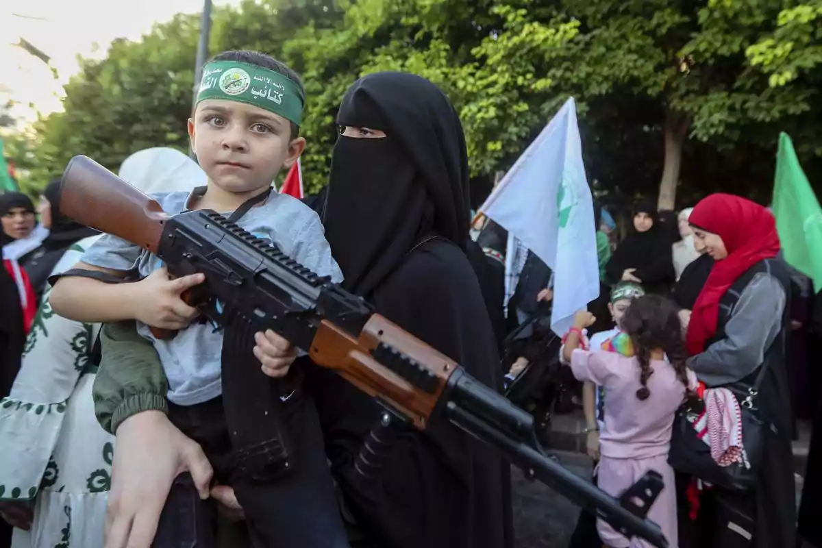 Un niño con una cinta en la cabeza sostiene un arma mientras está en brazos de una persona vestida de negro, rodeado de más personas y banderas en un entorno al aire libre.