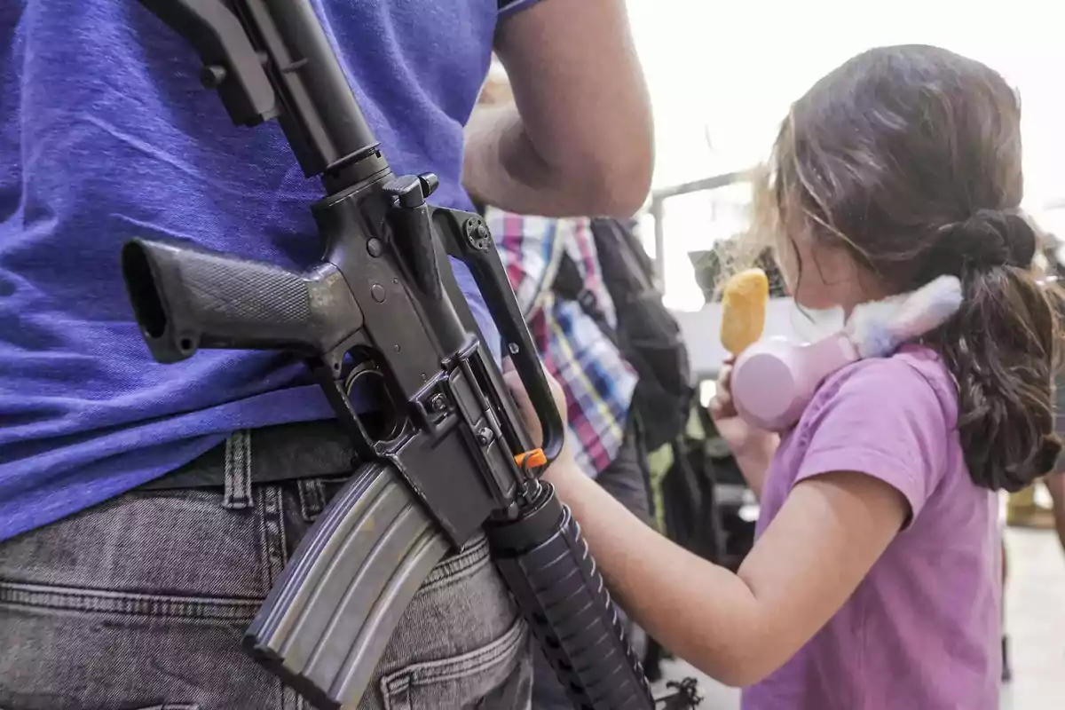 Un hombre con una camiseta azul lleva un rifle colgado mientras una niña con una camiseta rosa y auriculares come un helado.