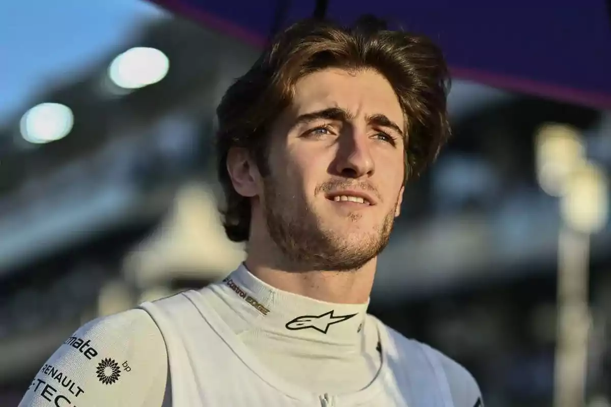 Un hombre joven con cabello castaño y barba ligera, vistiendo un traje de carreras blanco, mira hacia el horizonte con una expresión pensativa.