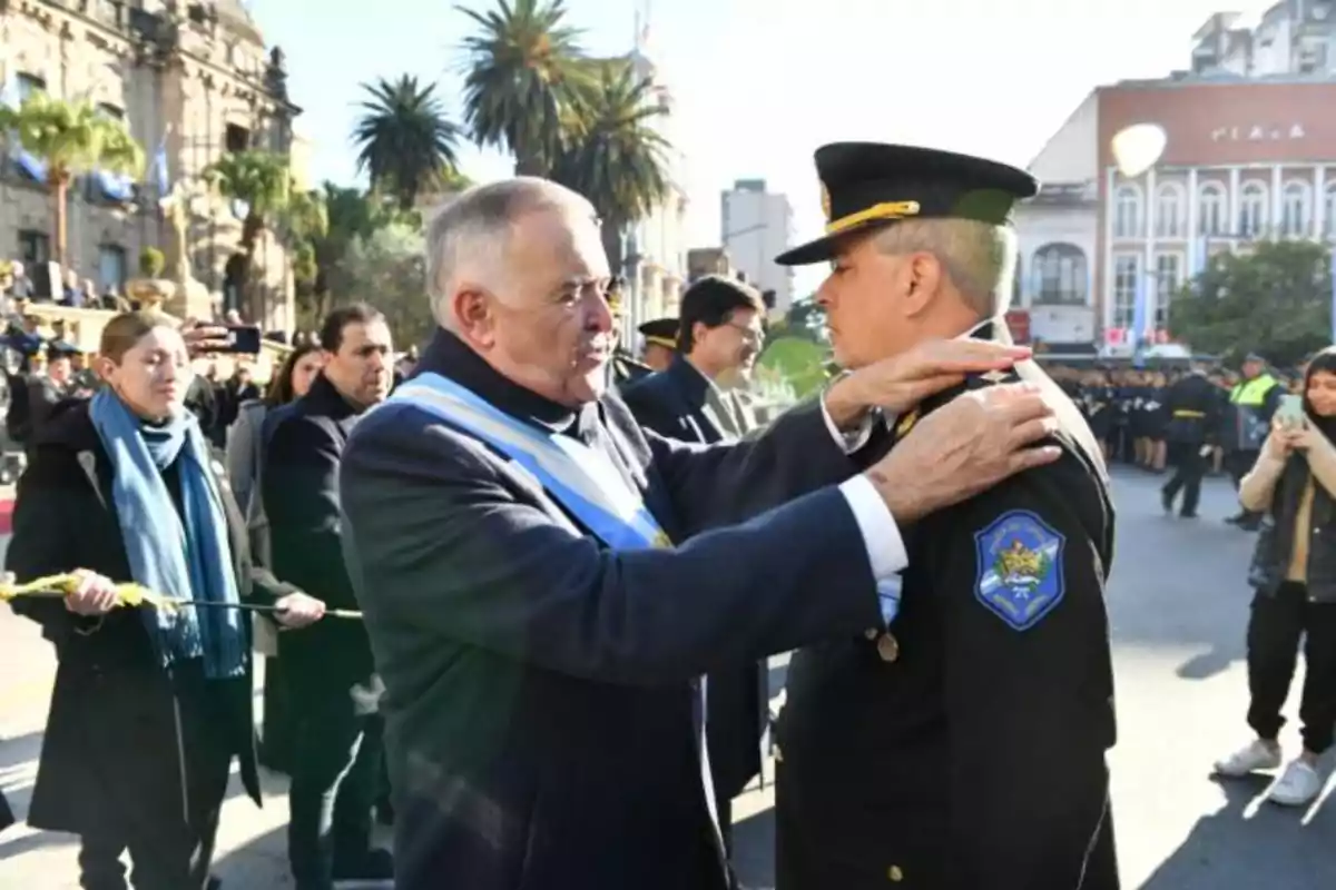 Un hombre mayor con una banda azul y blanca felicita a un oficial de policía en una ceremonia al aire libre con personas observando y edificios de fondo.