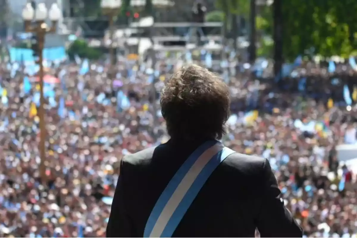 Una persona con una banda presidencial se dirige a una multitud en un evento al aire libre.