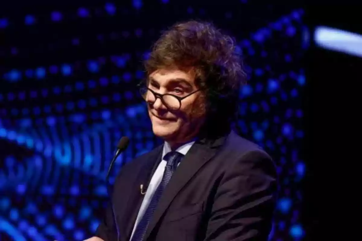 Un hombre con gafas y traje oscuro hablando en un micrófono con un fondo azul iluminado.