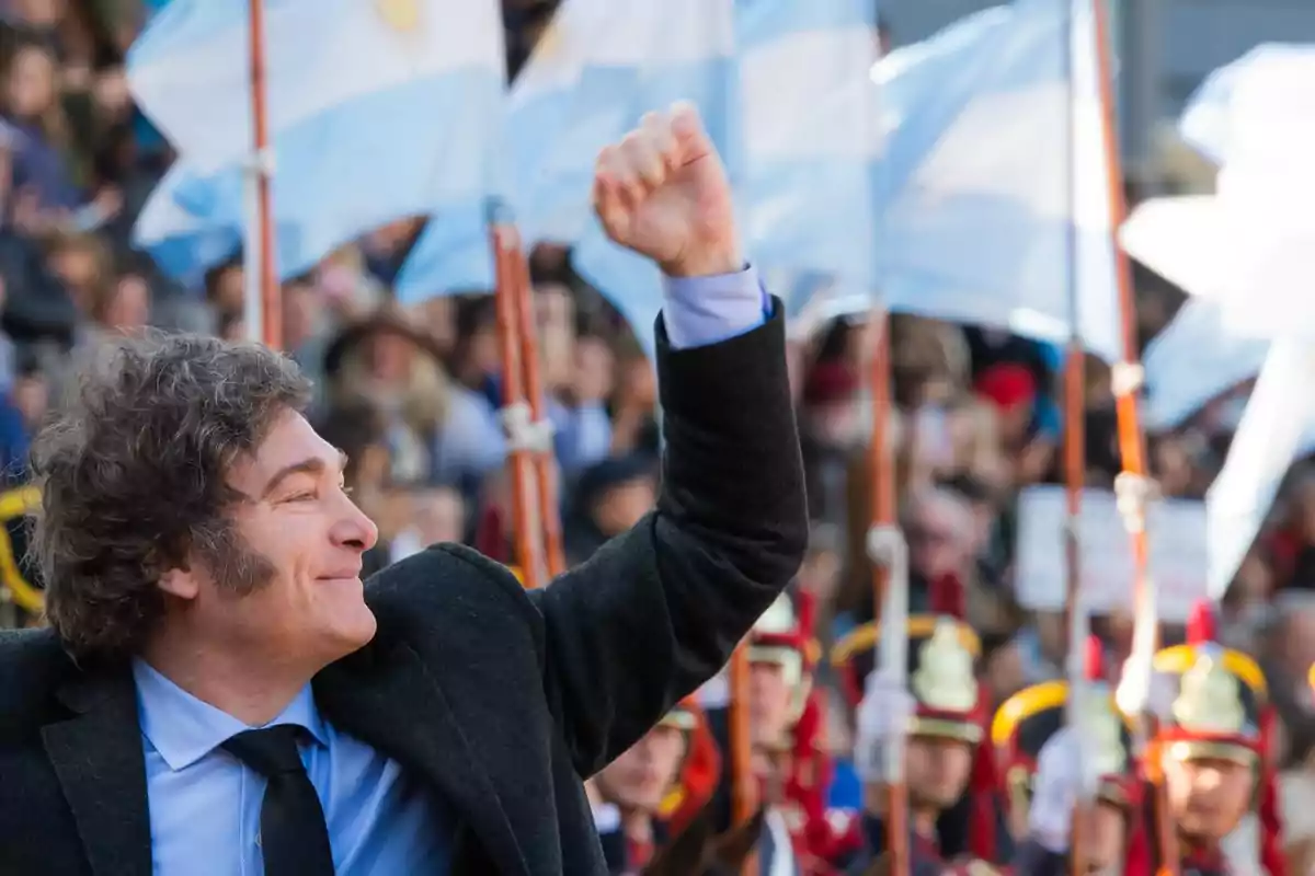 Un hombre sonriente levanta el puño en un evento público con banderas argentinas de fondo.