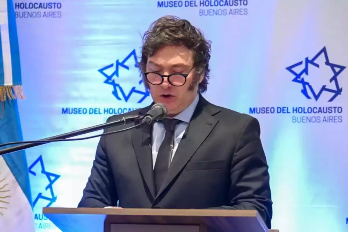 Un hombre con gafas y traje habla en un podio frente a un fondo del Museo del Holocausto de Buenos Aires.