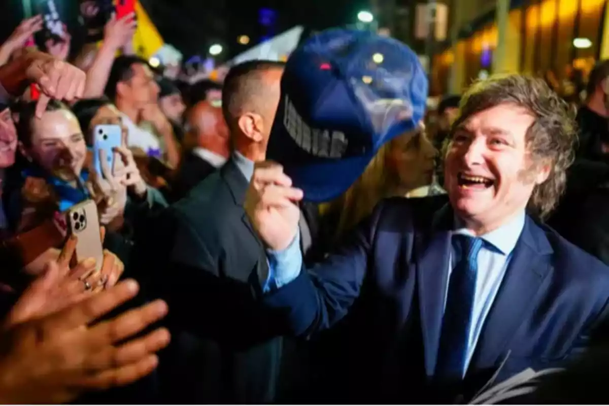 Una persona sonriente con traje y corbata sostiene una gorra azul mientras es rodeada por una multitud que toma fotos.