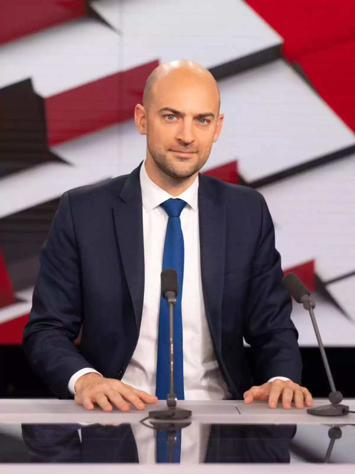 Un hombre calvo con traje oscuro y corbata azul está sentado detrás de un escritorio con micrófonos, frente a un fondo con diseño geométrico en rojo, blanco y negro.