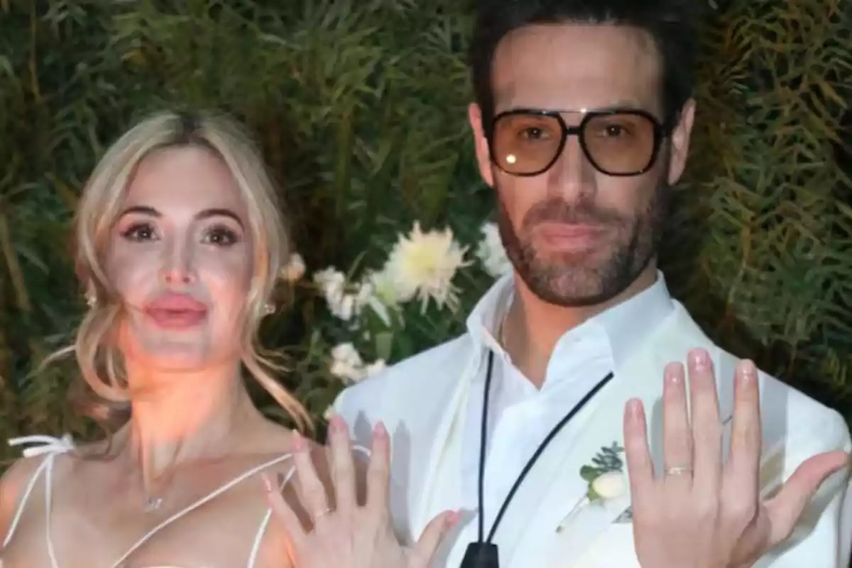 Una pareja vestida de blanco muestra sus anillos de boda frente a un fondo de vegetación.