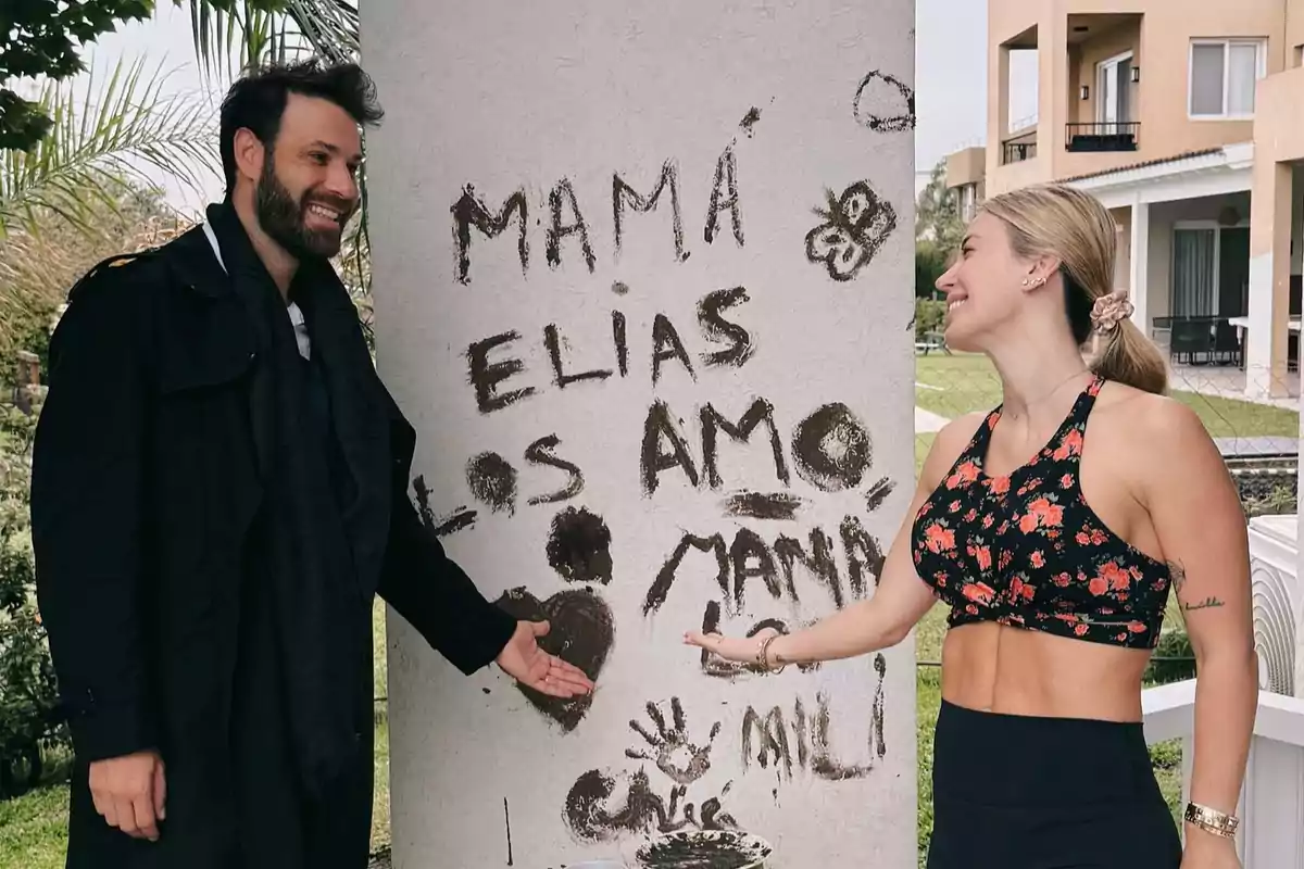 Una pareja sonríe mientras señala un mensaje escrito en una pared que dice "MAMÁ ELÍAS LOS AMO" junto a dibujos de una mariposa y un corazón.