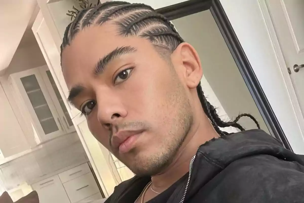 Un hombre con trenzas posando frente a un espejo en una habitación iluminada.