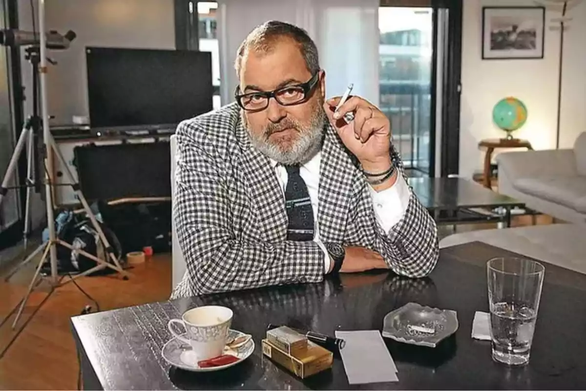 Un hombre con barba y gafas sostiene un cigarrillo mientras está sentado en una mesa con una taza de café, un cenicero y un vaso de agua en un ambiente interior.