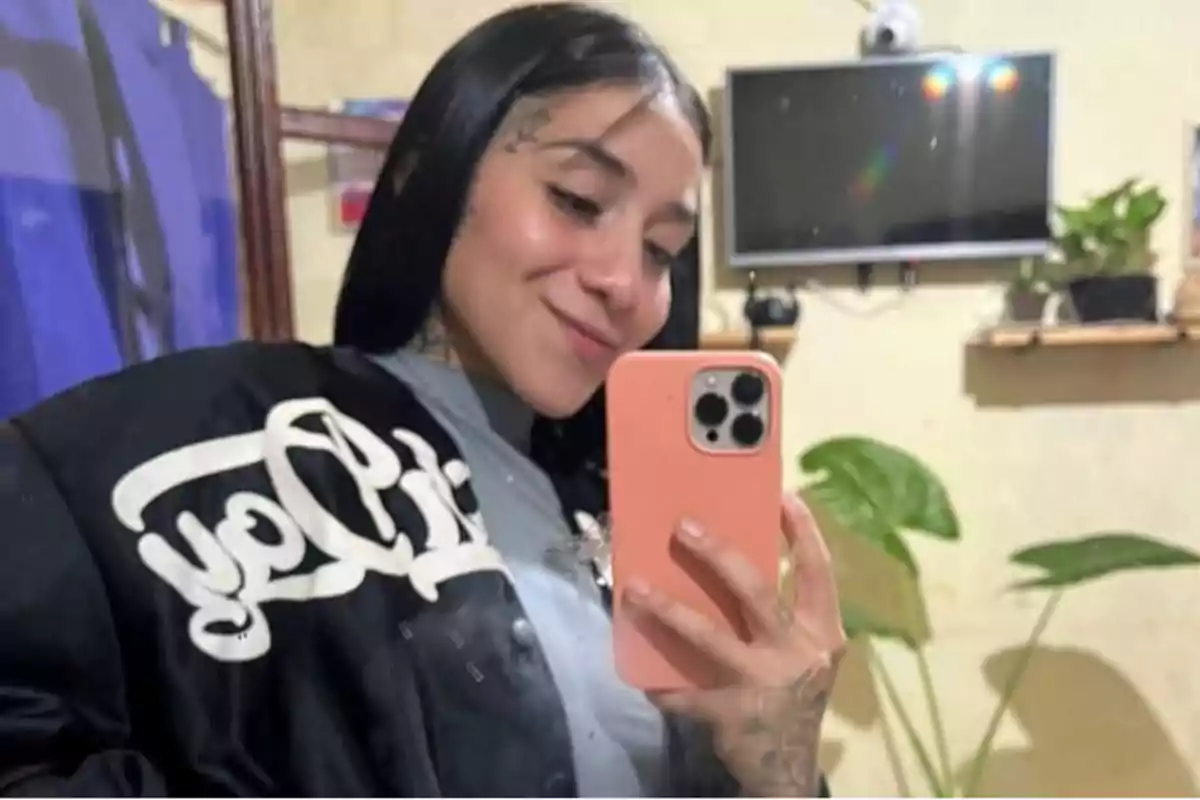 Una persona con cabello oscuro se toma una selfie frente a un espejo, sosteniendo un teléfono con funda rosa, lleva una chaqueta negra con letras blancas y hay una televisión y plantas en el fondo.