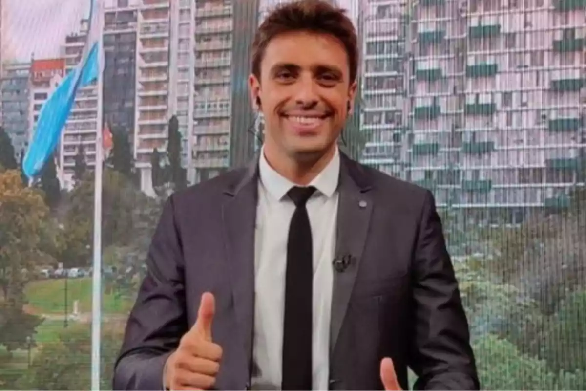 Un hombre sonriente con traje y corbata muestra un gesto de aprobación con el pulgar levantado, con un fondo de edificios y una bandera ondeando.