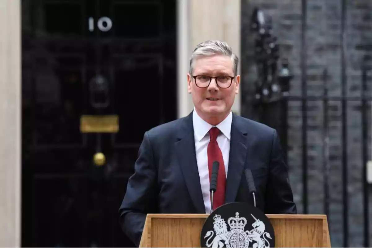 A man with glasses and a dark suit speaks in front of a lectern with the emblem of the United Kingdom, with the door of 10 Downing Street in the background.