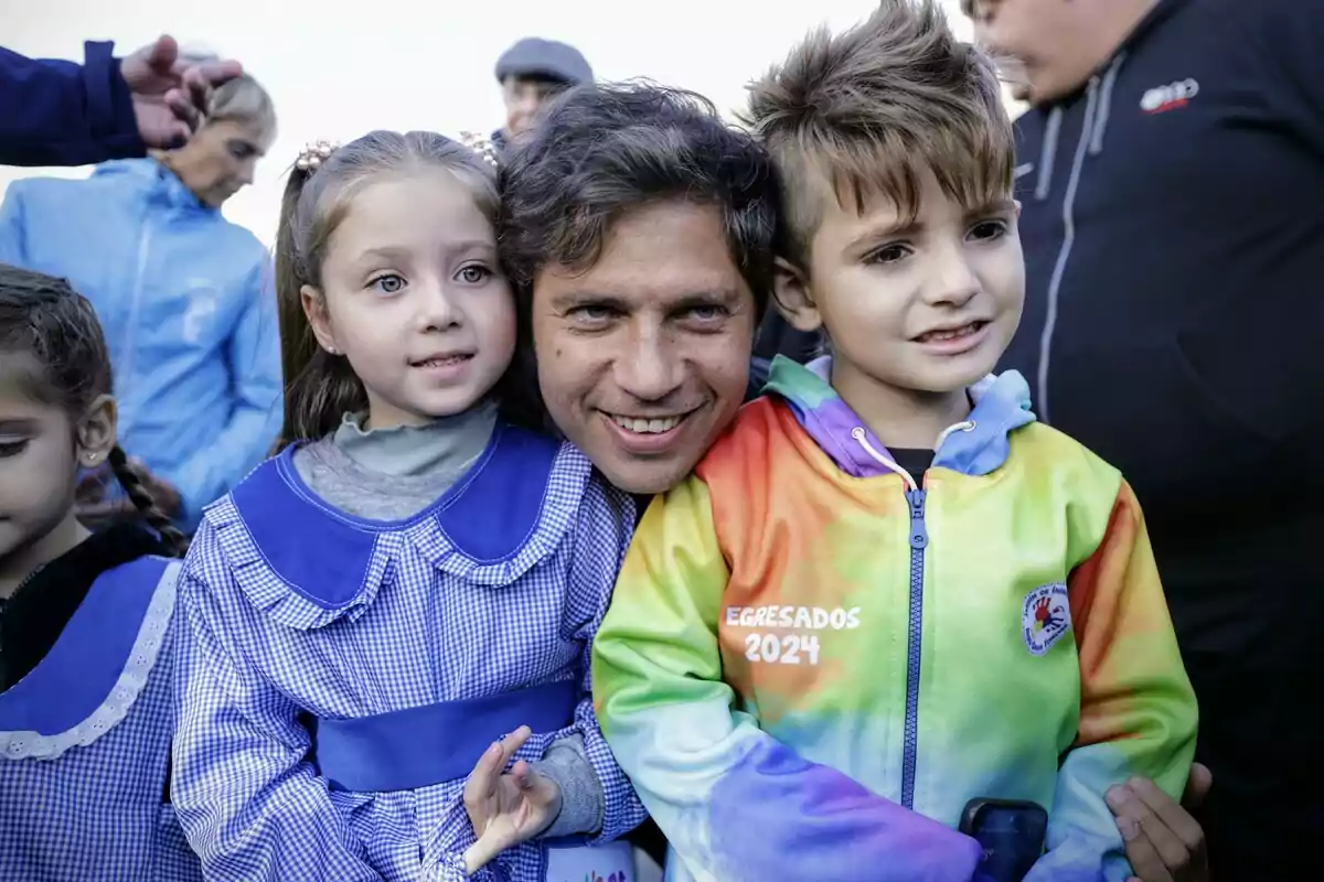 Un hombre sonriente abraza a dos niños pequeños, uno con uniforme escolar azul y otro con una chaqueta colorida que dice "Egresados 2024", mientras otras personas están en el fondo.