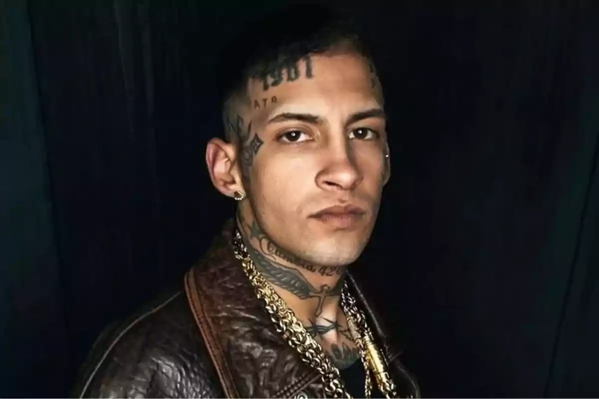 Un hombre con tatuajes en el rostro y el cuello, vistiendo una chaqueta de cuero y cadenas doradas.