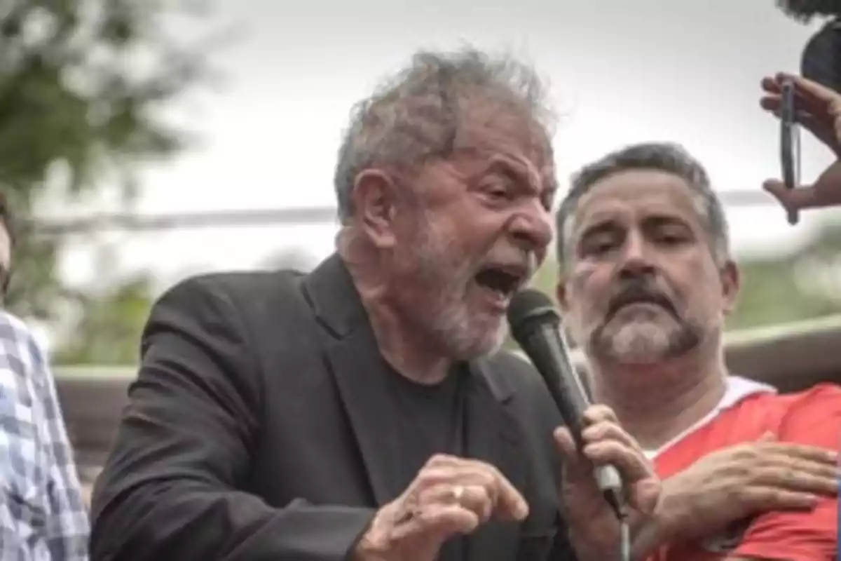 Un hombre mayor con barba hablando en un micrófono durante un evento al aire libre.