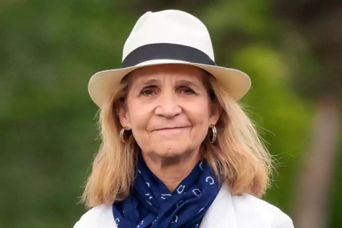 La infanta Elena de España con sombrero blanco y bufanda azul sonríe al aire libre.