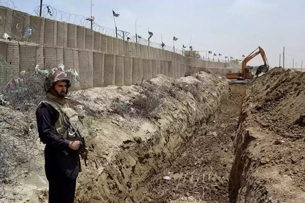 Un soldado armado vigila junto a una zanja en construcción cerca de una valla de alambre de púas, mientras una excavadora trabaja al fondo.