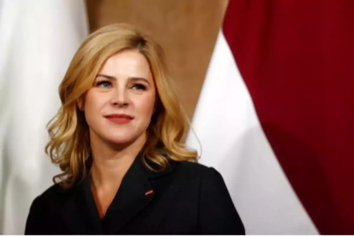 Una mujer rubia con chaqueta oscura frente a un fondo con colores rojo y blanco.