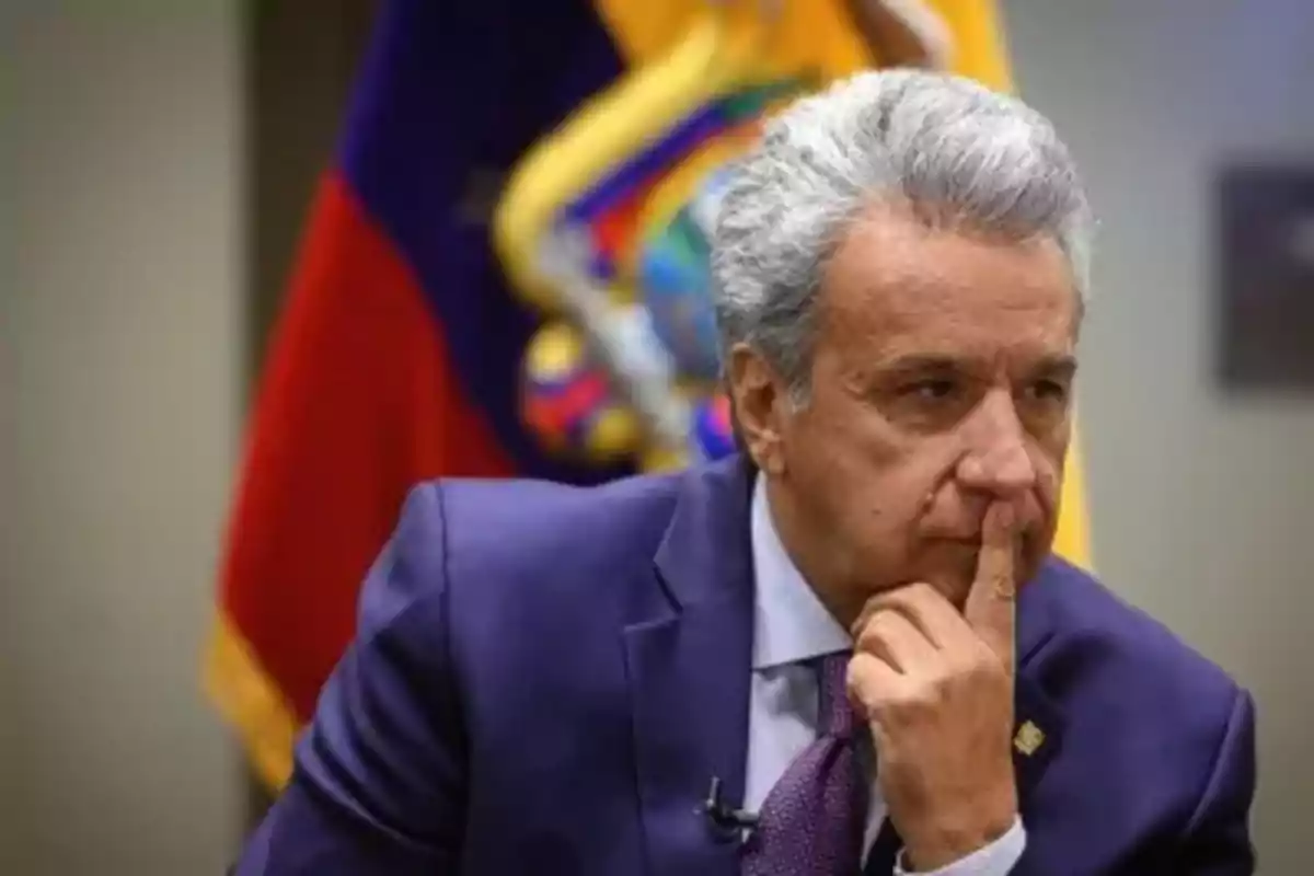 Un hombre de cabello canoso en traje azul con expresión pensativa y una bandera de fondo.