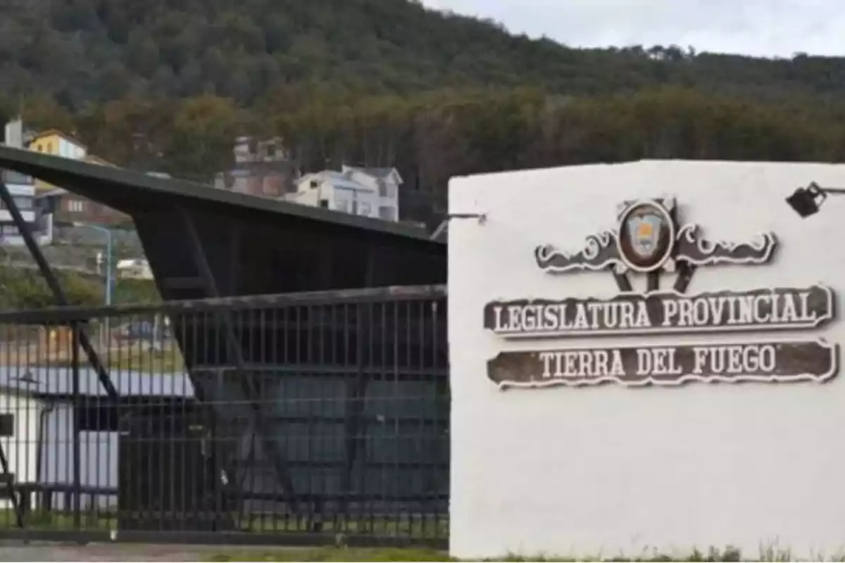 Entrada de la Legislatura Provincial de Tierra del Fuego con un cartel en un muro blanco y un fondo de colinas y casas.