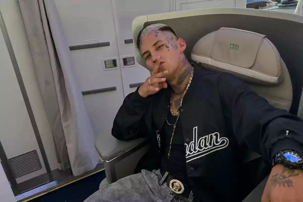 Un hombre con tatuajes en la cara y el cuello está sentado en un asiento de avión, vistiendo una chaqueta negra y un reloj azul.