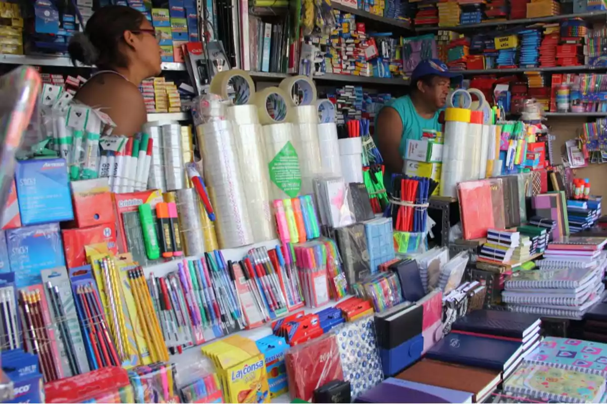 Una tienda de papelería con una amplia variedad de productos como cuadernos, lápices, bolígrafos, cintas adhesivas y otros artículos de oficina, atendida por dos personas detrás del mostrador.