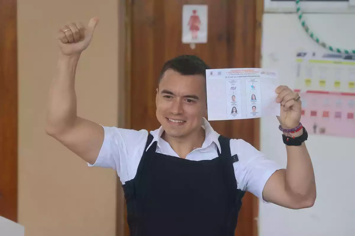 Un hombre sonriente levanta el pulgar y sostiene una papeleta de votación en un lugar de votación.