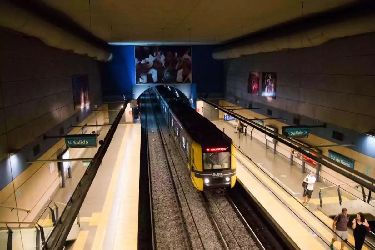 Un tren amarillo del metro se aproxima a una estación subterránea con andenes a ambos lados y señales de salida visibles.