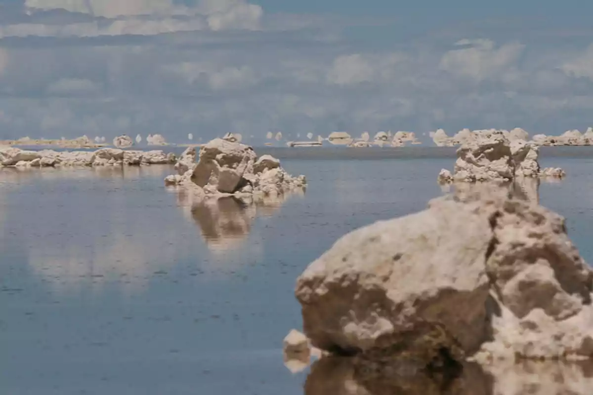 A view of a desert landscape with white rock formations reflected on a calm water surface under a partly cloudy sky.