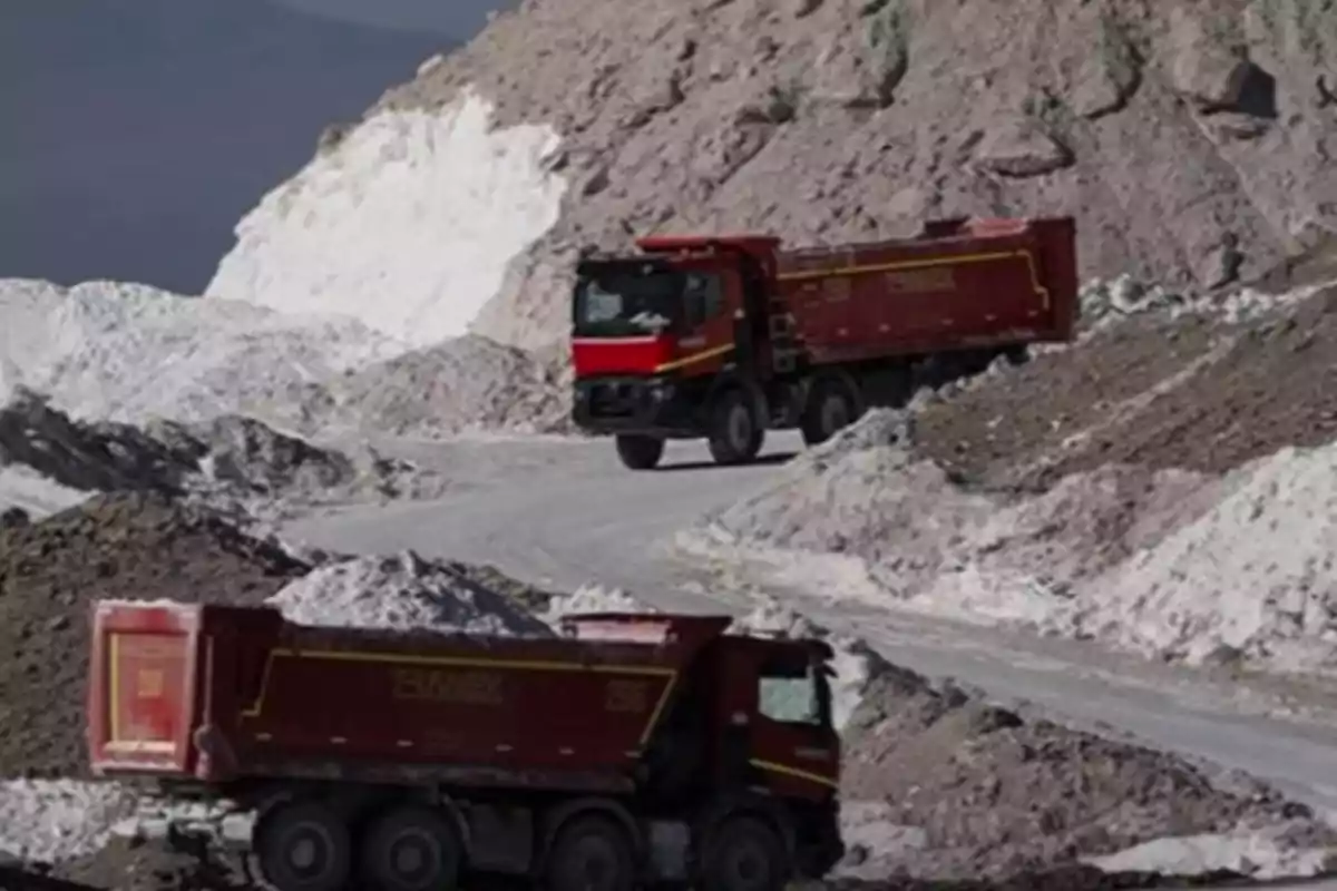 Two red trucks transport material on a road surrounded by mountains of dirt and rocks.