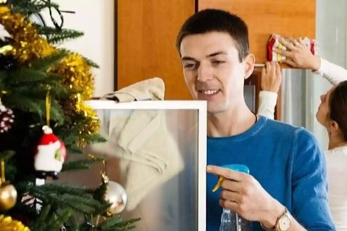 Hombre limpiando un espejo cerca de un árbol de Navidad decorado mientras una mujer limpia un mueble al fondo. tradición navideña en Lituania.