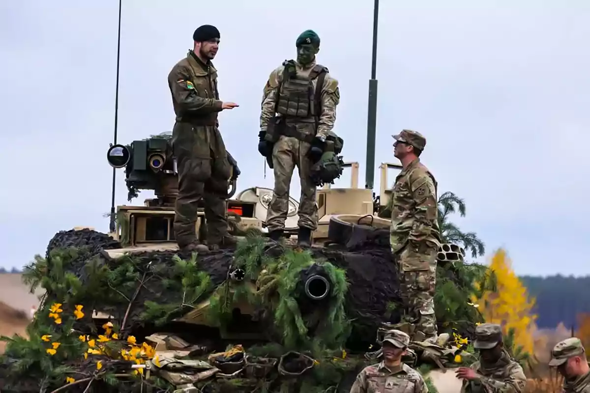 Soldados de diferentes nacionalidades conversan sobre un tanque camuflado con vegetación en un entorno al aire libre.