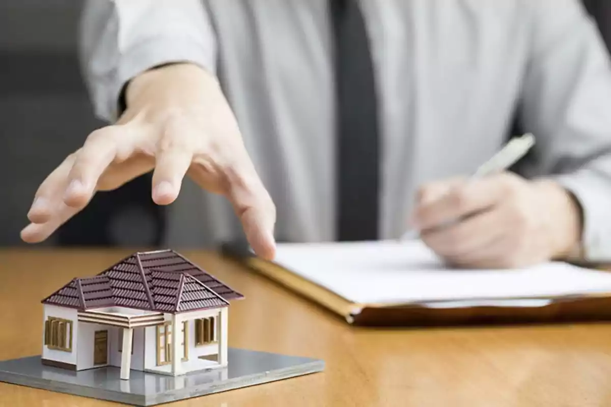 A hand extended over a model house while a person writes on a document.