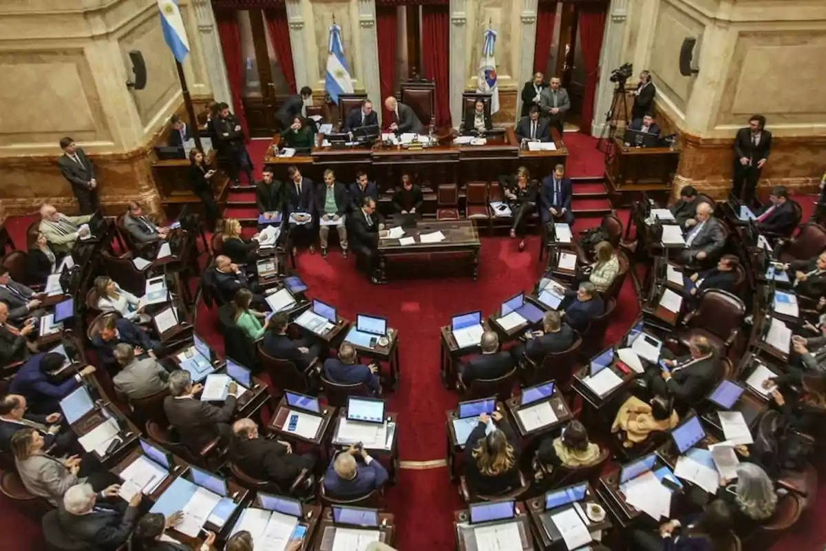 Vista aérea de una sesión parlamentaria en un recinto con banderas argentinas y personas trabajando en escritorios con computadoras portátiles.