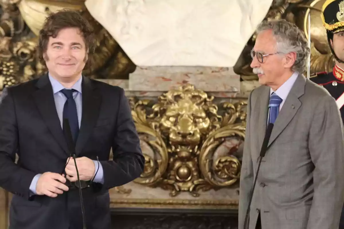 Dos hombres de traje en un evento formal con un fondo decorativo y un guardia detrás.