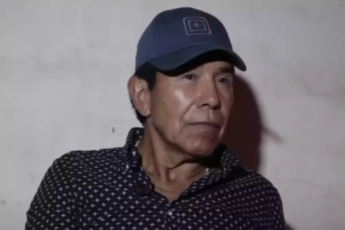 Un hombre con gorra azul y camisa de estampado geométrico en un fondo claro.